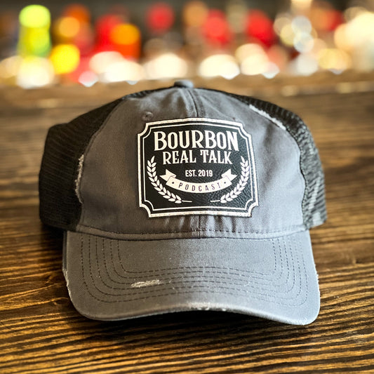 Distressed Unstructured Trucker Hat with leather patch on wooden counter top with whiskey bottles in the background