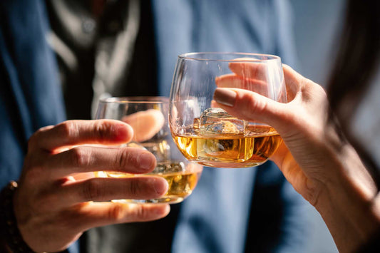 image of two hands clinking drinking glasses with bourbon over ice 