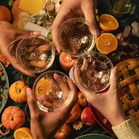 Image of 4 hands holding glasses of a cocktail over a full table of fall foods for thanksgiving