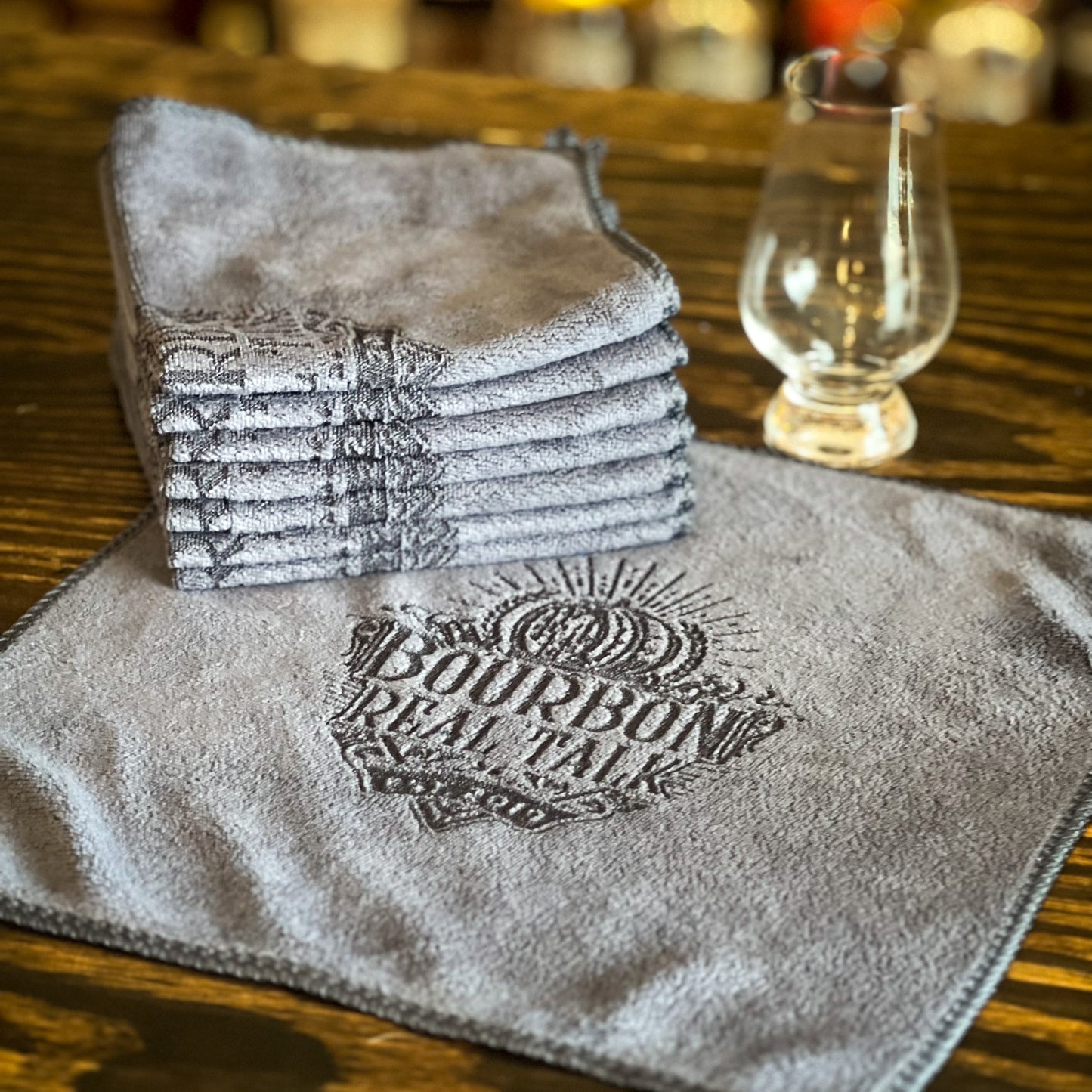 gray microfiber cloth with laser printed logo, there is a stack of folded cloths  next to a glencairn whiskey glass