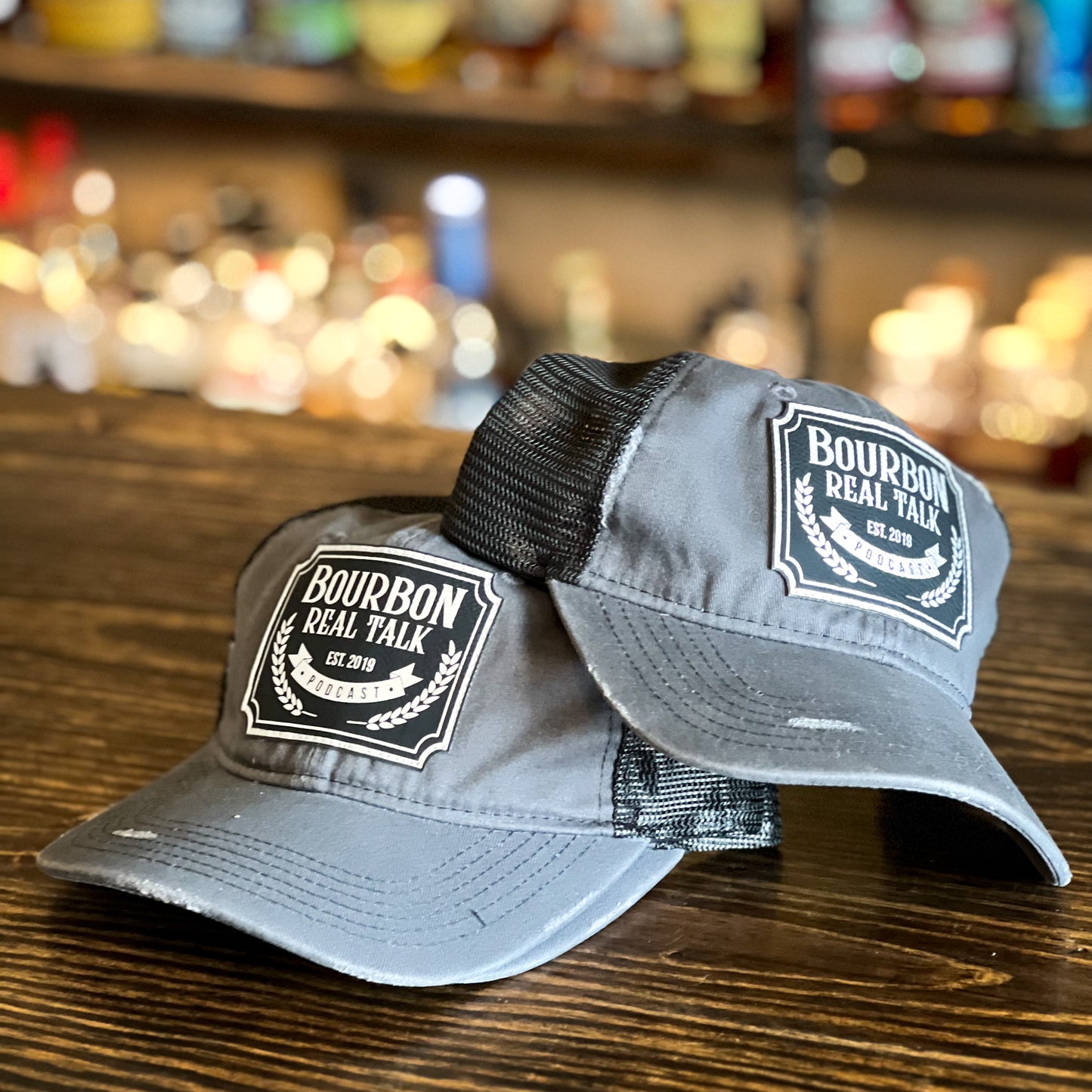 Distressed Unstructured Trucker Hat with leather patch, photo  has two hats on wooden counter top with whiskey bottles  in the background