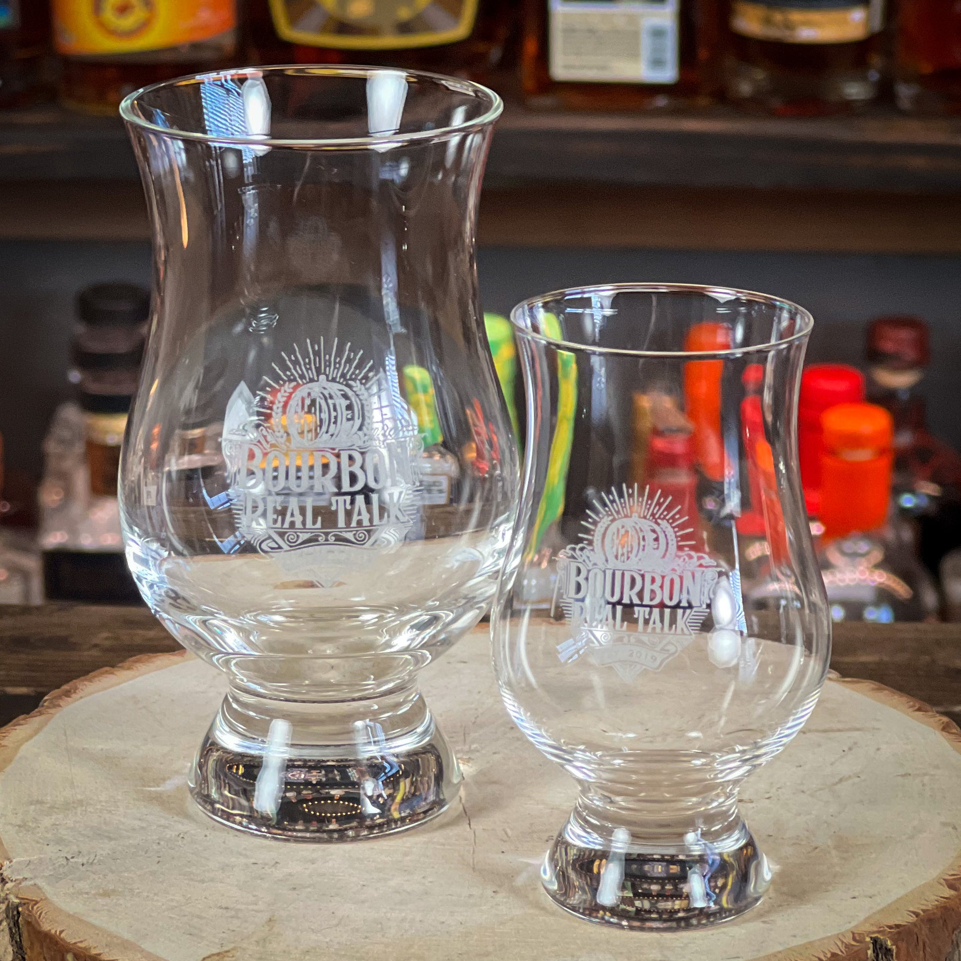 This photo shows two whiskey nosing glasses nosing glasses side by side, one 6oz. one 3oz. on a wooden block with whiskey bottles in the background.