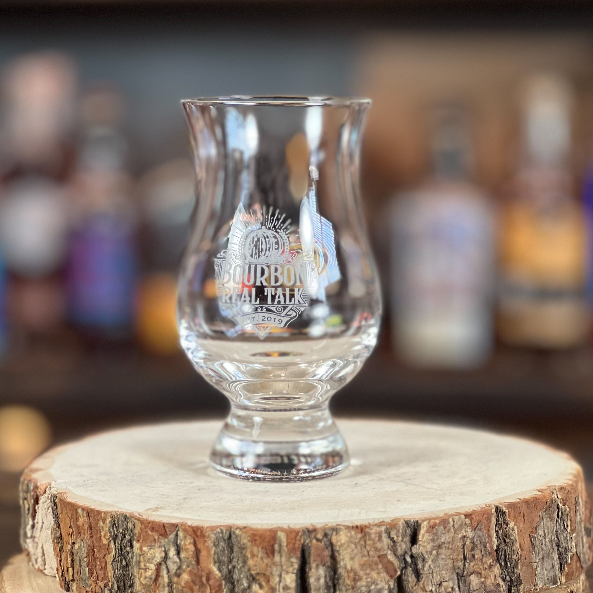 This photo shows one whiskey tasting nosing glass  on a wooden block with whiskey bottles in the background.