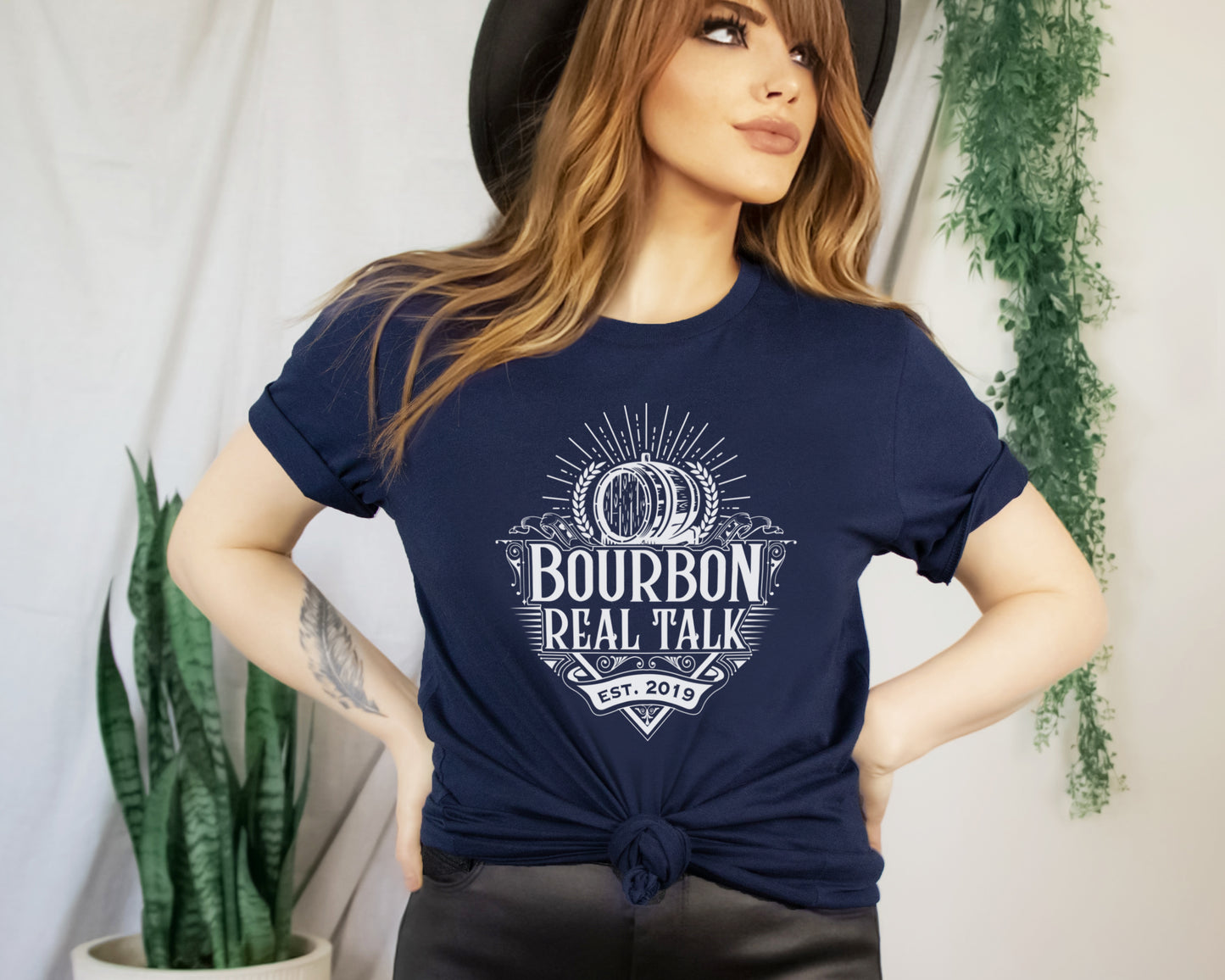 woman with red hair, black hat, black leather pants and a navy Bourbon Real Talk shirt, tied in the front