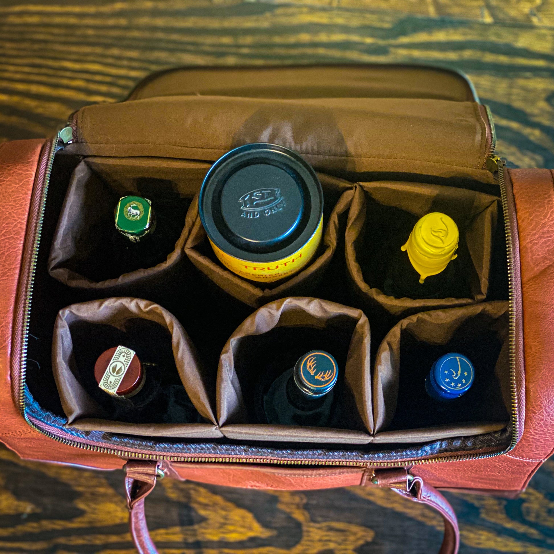 Front of the 6 Bottle Whiskey Carrier, with front pocket, zipper around the top, shoulder strap and handles, shows the 6 removable velcro compartments  filled with various sizes and shapes of whiskey bottles